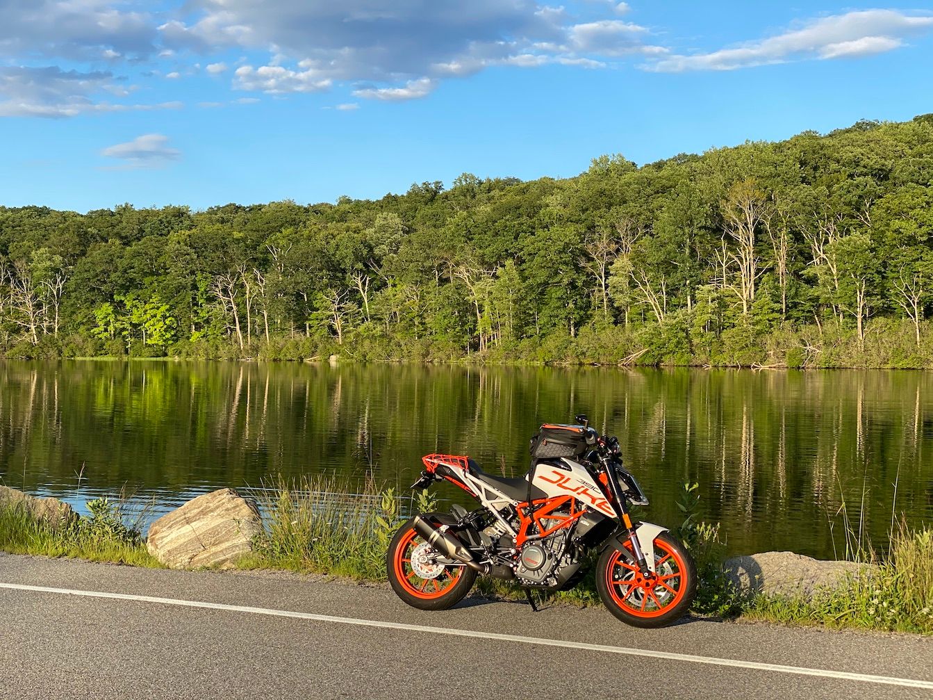 My Duke 390 sitting on the side of the road with a background of a pond and some woods reflecting on it.