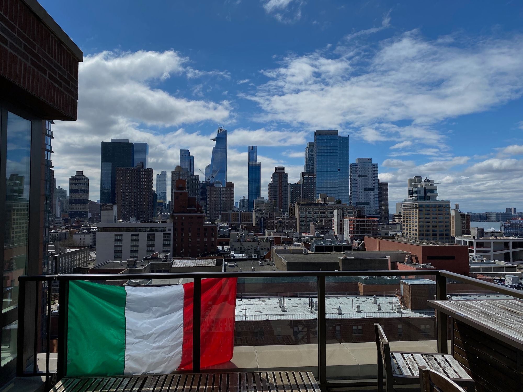 In the background, a skyline with six skyscrapers against a blue sky with a few bright clouds. In the foreground, a terrace with outdoor chairs and an Italian flag hung on the railing.