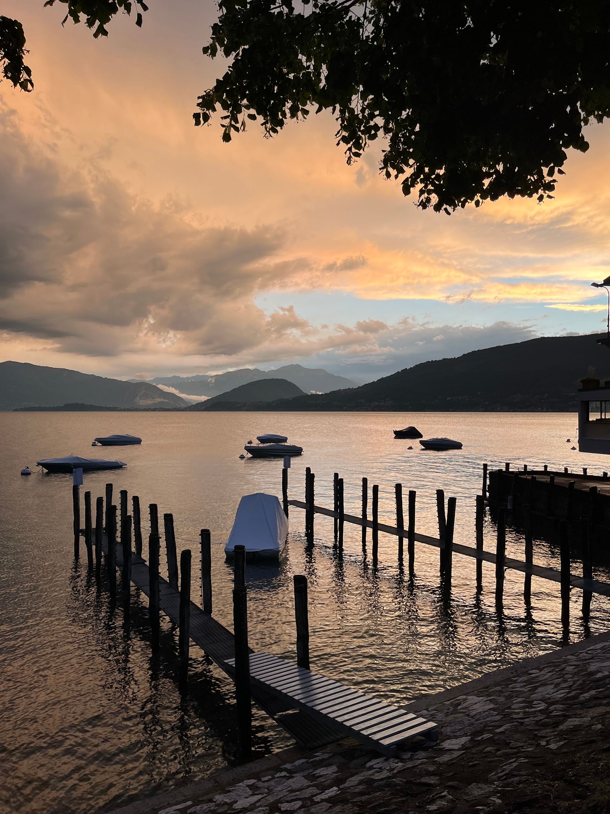 A picture of a lake taken from the shore. The sky is full of clouds colored orange from a recent storm. They reflect on the water, where a few boats sit anchored. Two wooden piers extend from the shore from right to left. In the distance mountains on the other side of the lake, and a small sliver of blue sky.