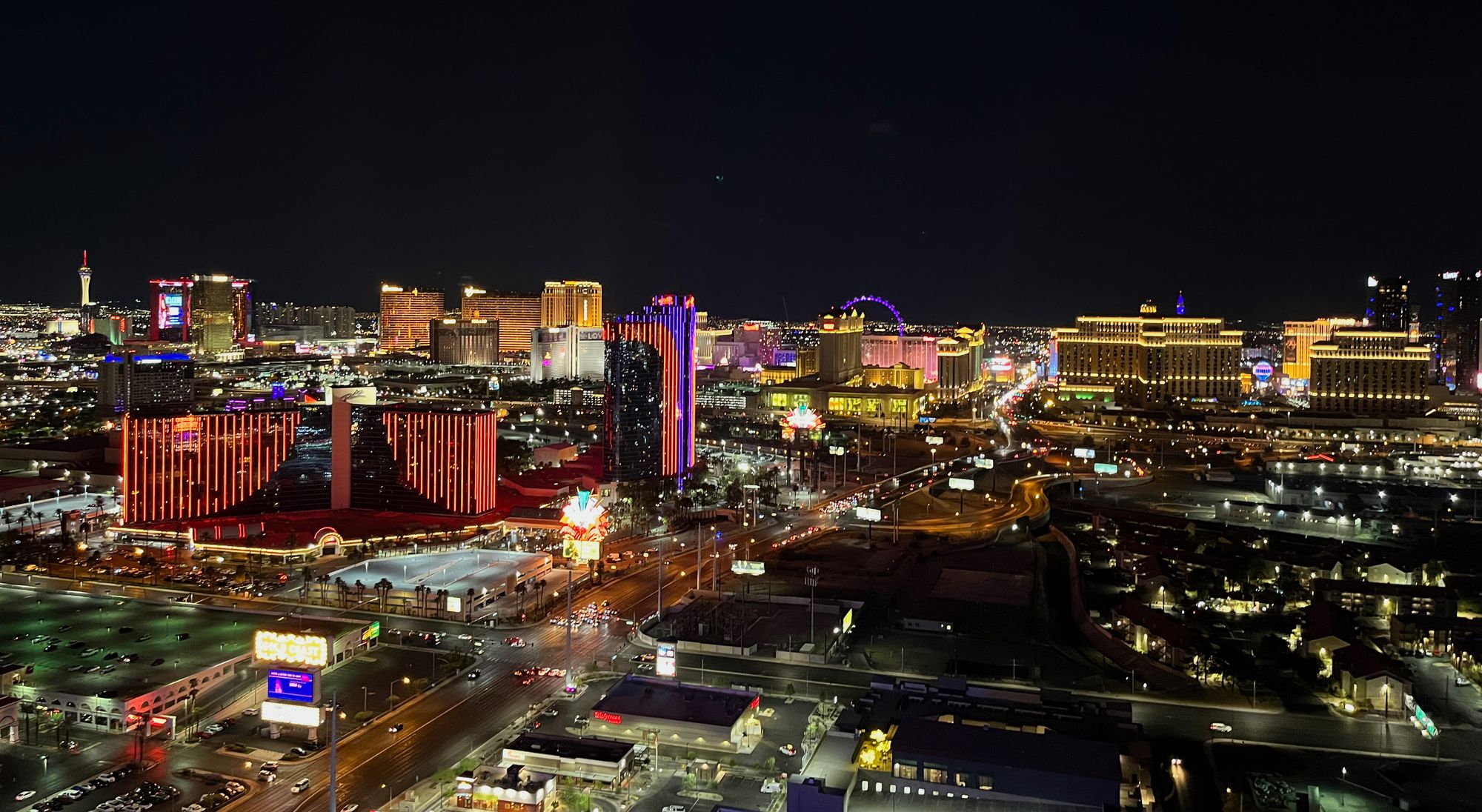 A night-time picture of the Las Vegas skyline taken from a high floor. In the foreground a large road, mostly empty, and two blocks of low buildings and empty space with a couple billboards with bright lights. Mid-frame, a string of high-rise casinos covered in sparkly lights. In the background, pitch black sky.