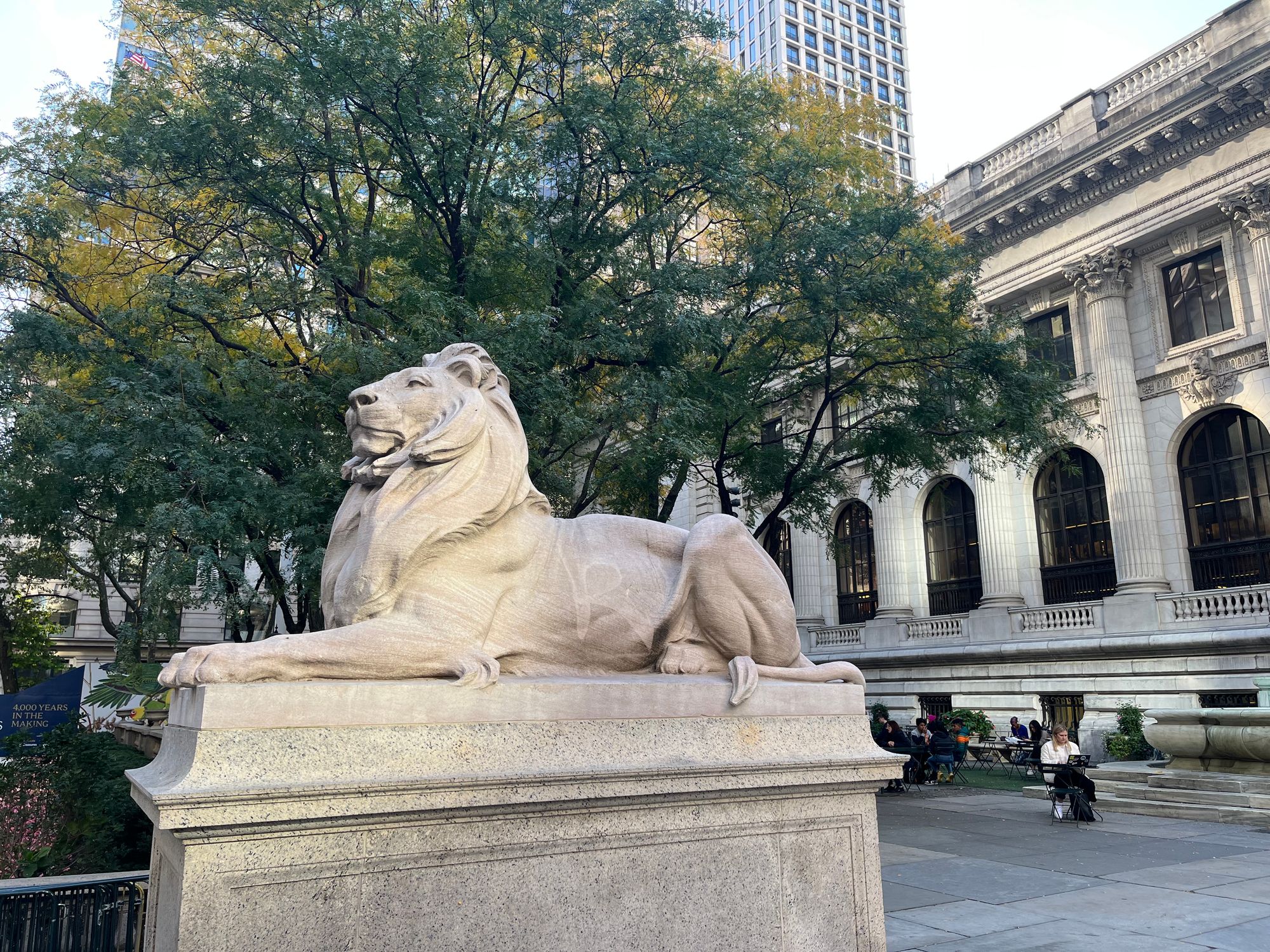 A large stone lion, bigger than life, on a pedestal pictured from the right. In the background, a tree, a building with white columns, and a skyscraper.