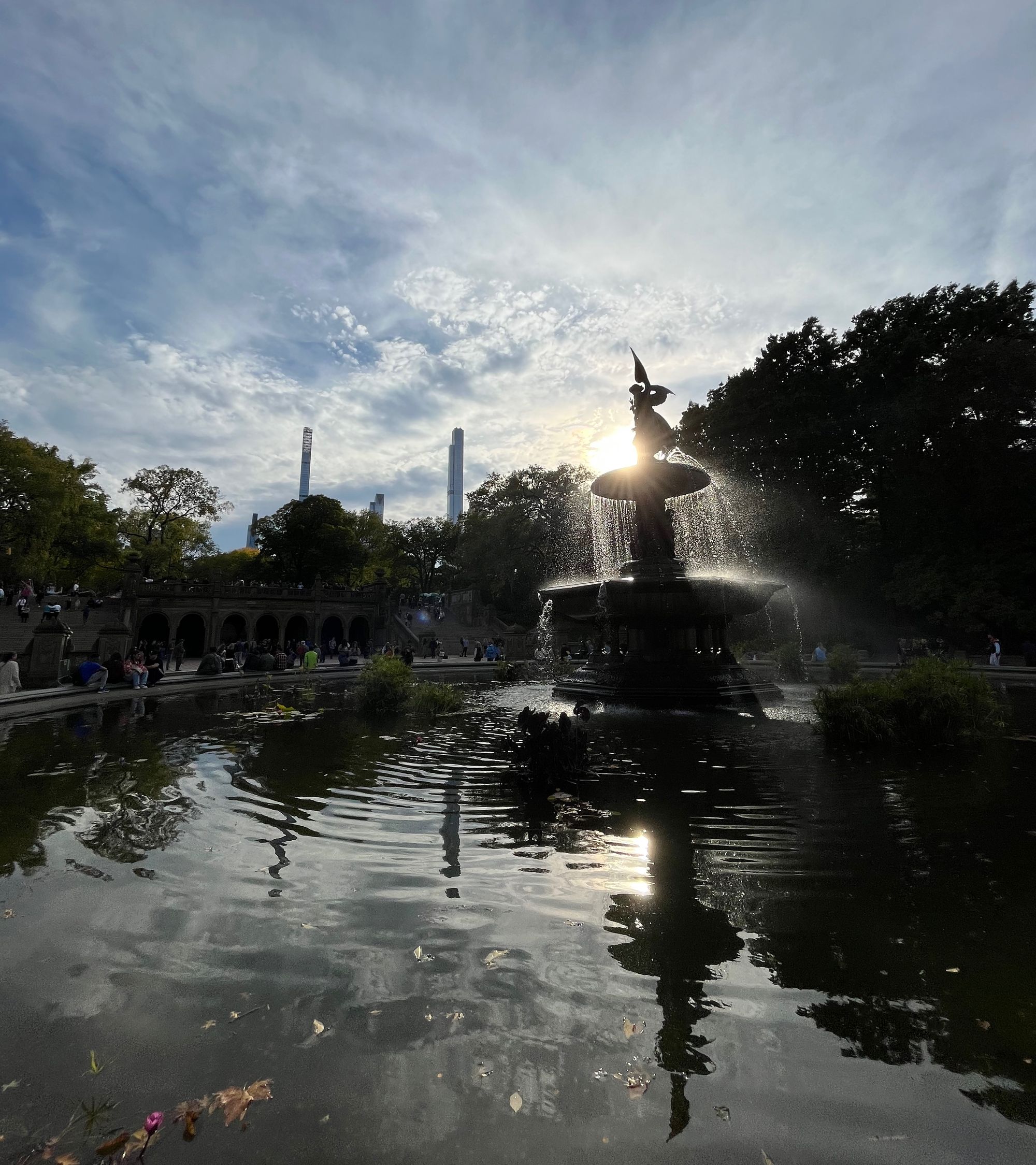 The Central Park fountain with the sun shining behind it and lighting up the water. In the distance, trees and a couple of the tall square skyscrapers. The bottom half is filled by the reflection of the fountain in the water.