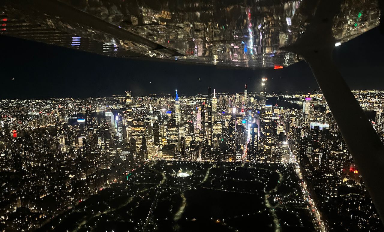 The New York skyline at night from a point above Central Park. A wing and a strut are visible at the top of the picture, and the city lights reflect off it.