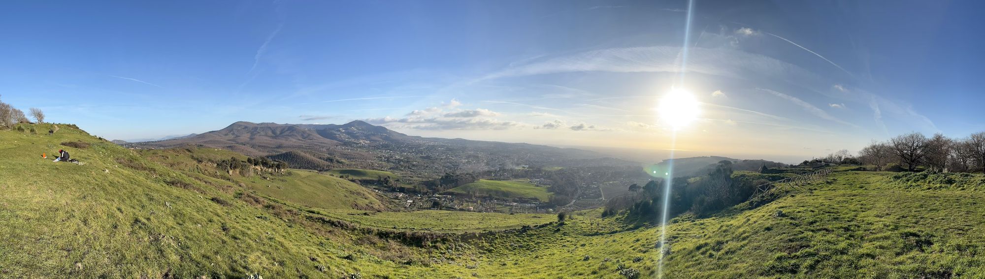 A panorama picture taken from a green hill. In the center the valley below is brown and dotted with houses. The sun is starting to set towards the right of the picture, in a smoky but clear light blue sky.