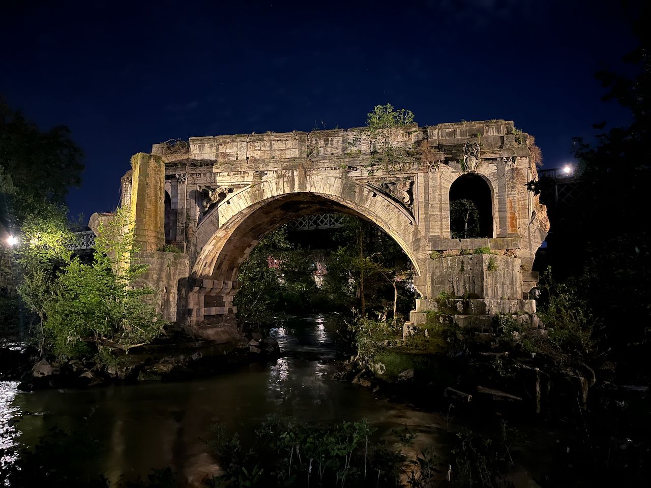 A single arch of a stone bridge is lit in the foreground, pictured from the side and below. The stone is greyed by rain and sediment, there's vegatation hiding the sides. The water passes under and around it. The night sky behind it is dark blue.