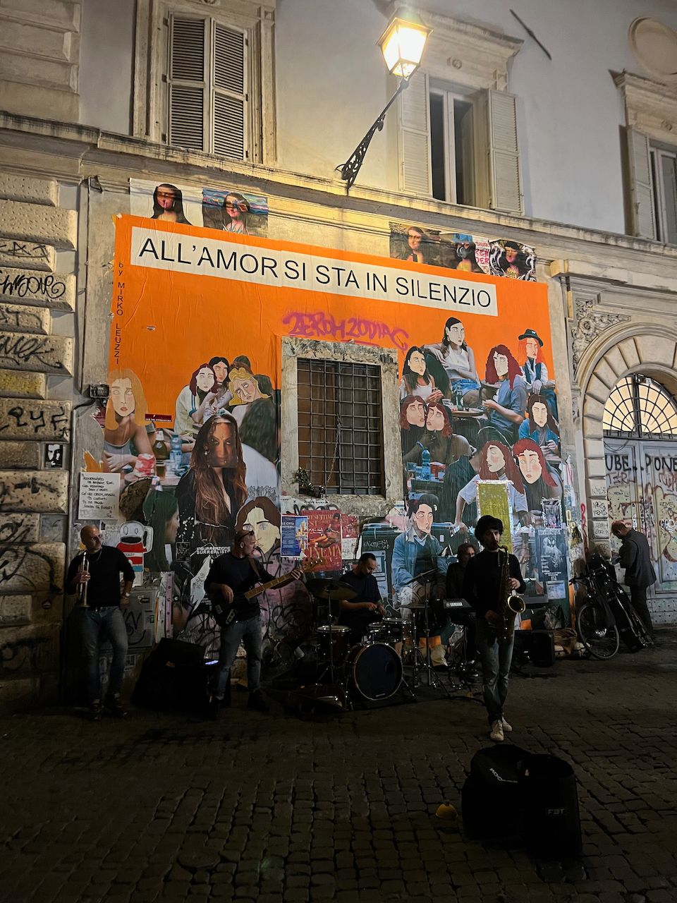A jazz band at night on a cobblestone road, against a wall with a large orange mural with drawn faces and the text "ALL'AMOR SI STA IN SILENZIO", below a street light. There's tenor sax, trumpet, bass, drums, and keyboard.
