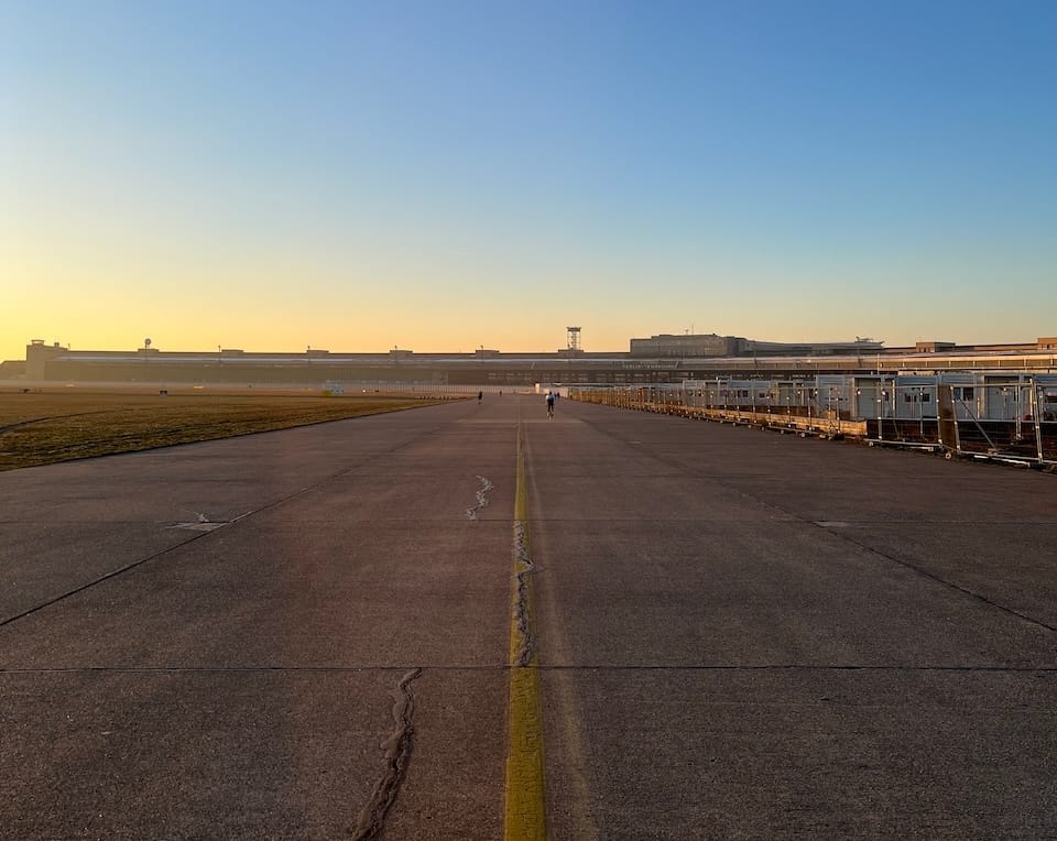 A cement taxiway pictured at sunset, from the middle of the yellow centreline. The airport terminal is visible on the horizon, and a patch of grass on the left.