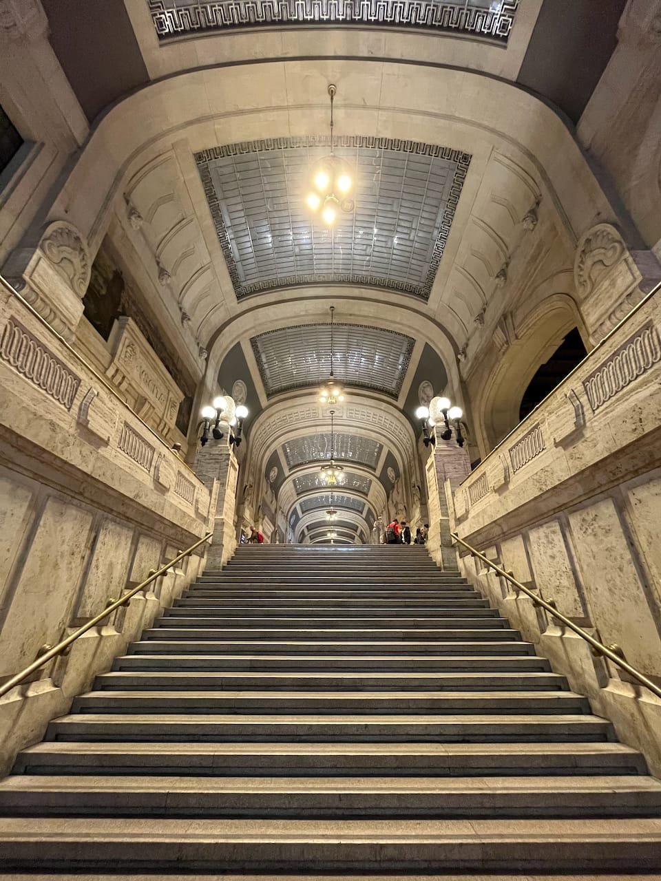A vertical shot of a stairwell from the bottom. Everything is white marble. Golden handrails. At the top of the stairs just the roof is visible: tall arches with glass portions and hanging chandeliers.