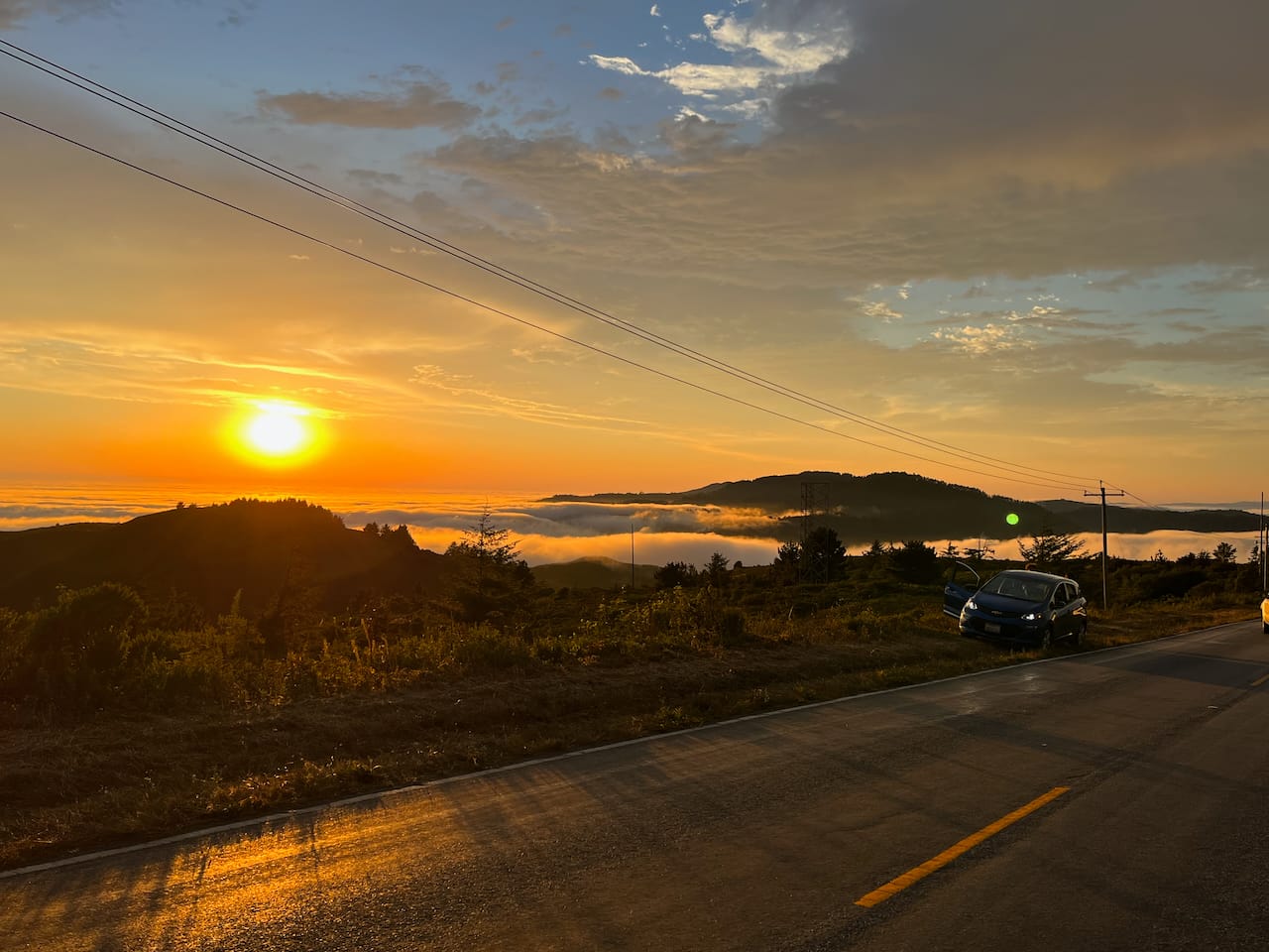 A fiery orange sunset, pictured from the side of the road. The valley below is filled with a sea of white clouds.