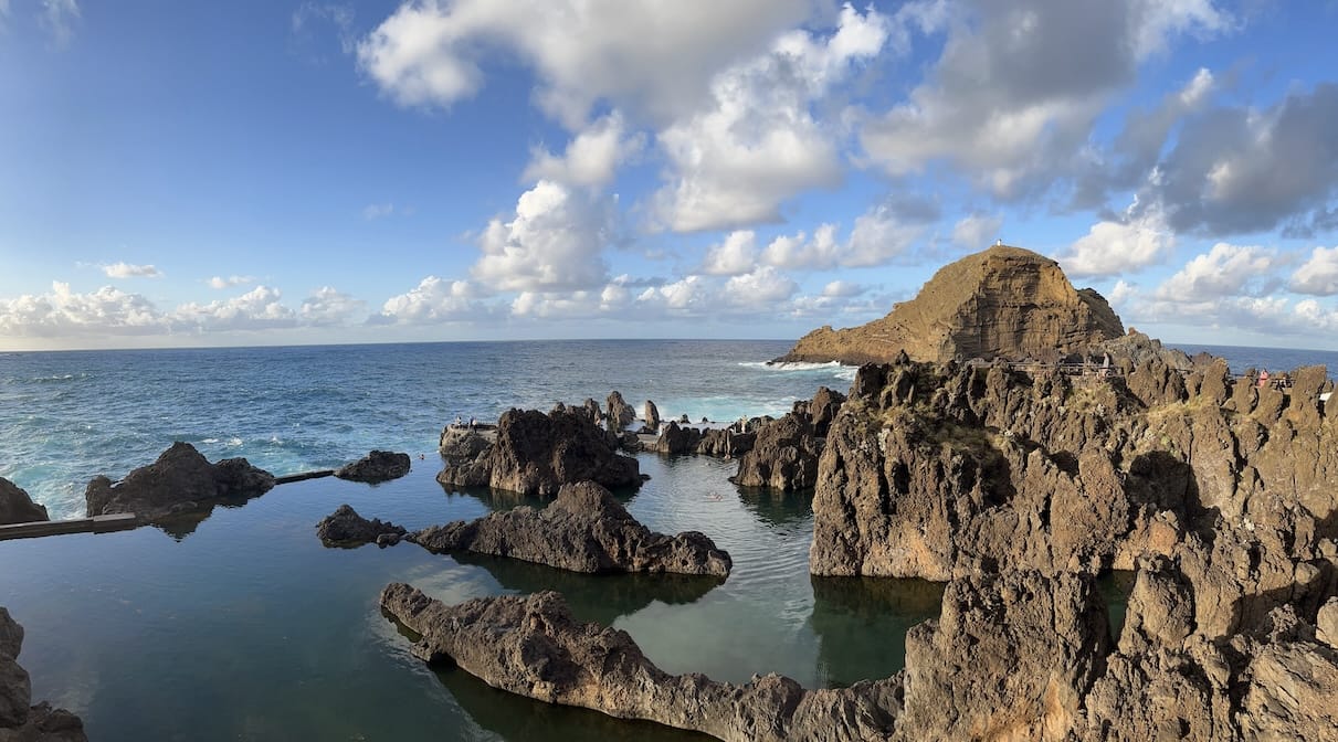 A natural pool with clear blue water, surrounded by dark volcanic rocks in the sunset light. The ocean is visible in the background, a few white clouds reflect on it. A rocky island has a lighthouse on the top.