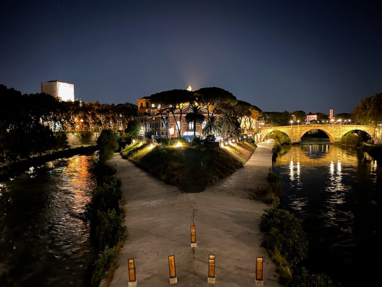 A nighttime view of Tiber Island in the middle of the Tiber River, featuring a lit stone bridge connecting the island, historic buildings with warm lighting framed by trees, and reflections of lights on the calm black water to the two sides.